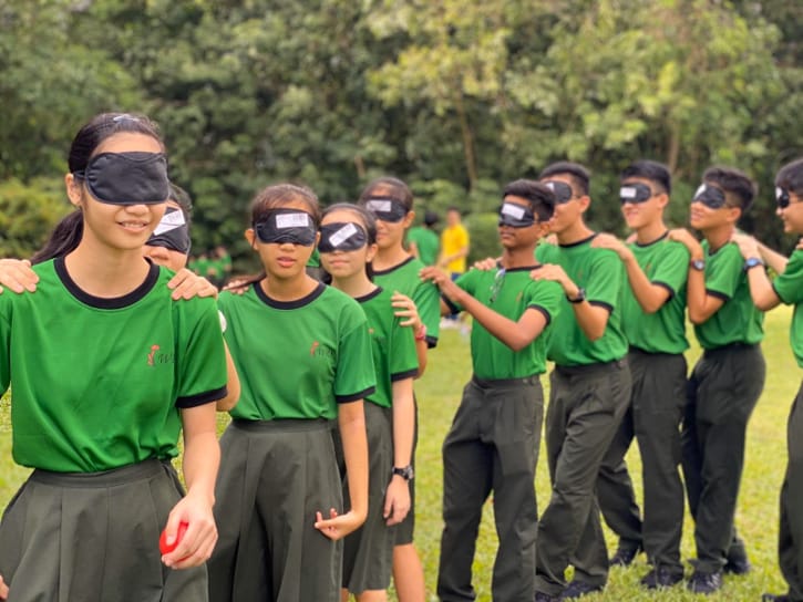 Students in green uniforms wear blindfolds and form a line, each with hands on the shoulders of the person ahead.